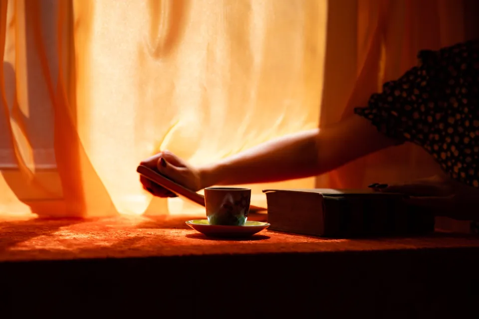 A hand opening the curtains, a cup and a book on the windowsill